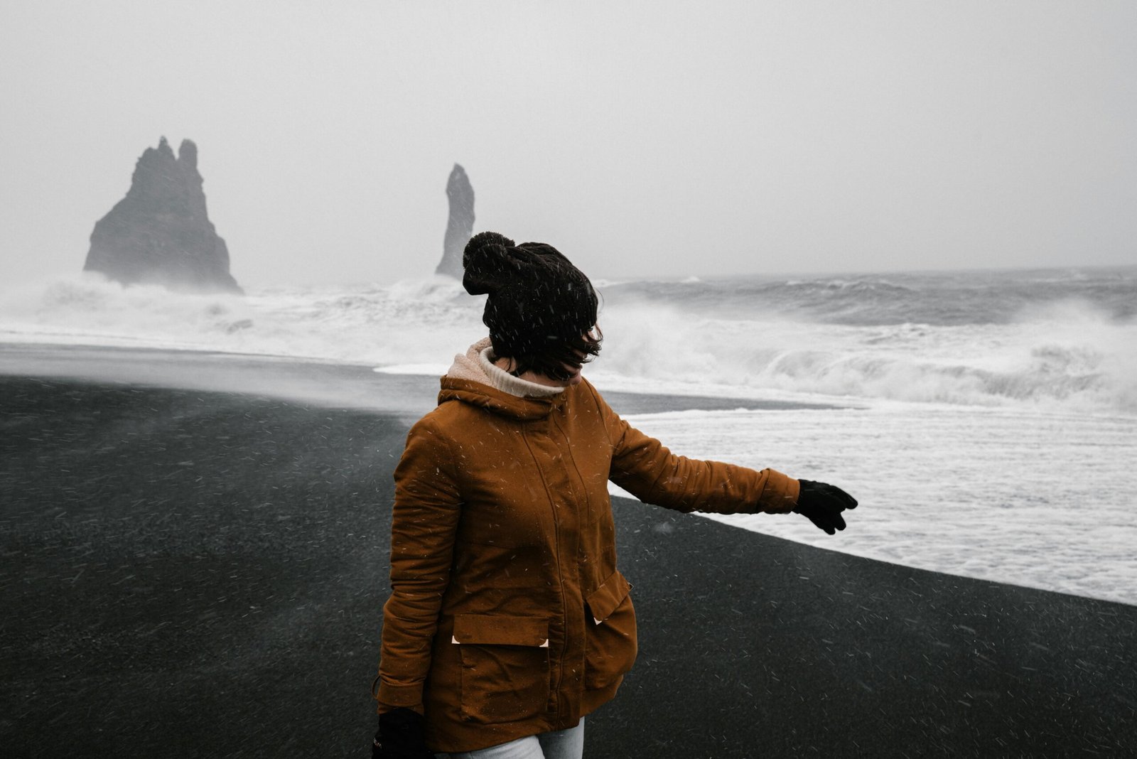 A woman in a brown jacket stands on a black sand beach in Iceland amidst a snowstorm.